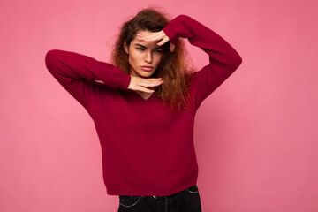 Portrait of young serious beautiful brunette curly woman with sincere emotions wearing trendy pink pullover isolated on pink background with copy space and grimacing with hands