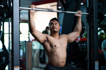 Fototapeta na wymiar Shirtless adult Asian men sweating while lift up the barbell workout machine for muscle part inside of fitness gym. Bodybuilding athlete sport training for body strength and good health.