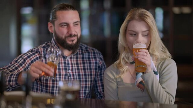 Front view positive man and woman toasting looking at camera drinking beer sitting at bar counter. Portrait of Caucasian friends or couple enjoying weekend leisure posing in pub indoors with alcohol