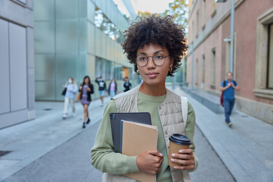 Outdoor shot of pensive curly Afro American student going to university drinks takeaway coffee holds tablet and notebooks wears round spectacles cassual clothes poses at street in publicity area