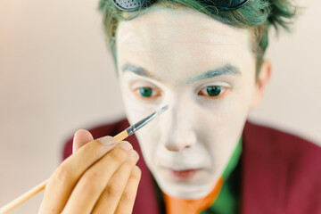 Mim is preparing makeup for performance. Clown cosplayer concept. Man with green hair in burgundy suit. Theatre atmosphere. Male actor is covering his face by white greasepaint using brush.