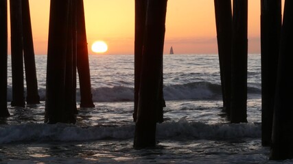 Waves splashing under pier, sunset in Oceanside, California USA. Ocean water, sun and wooden piles. Pillars from wood and seascape below boardwalk. Pacific beach resort romantic twilight atmosphere.