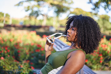 a dark-skinned girl with a lush curly black hairstyle in the hands of the girl with white coloured glasses in a park with flowers outdoors.