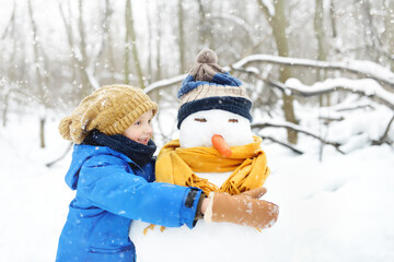 Little boy building snowman in snowy park. Child embracing snowman wearing hat and scarf. Active outdoors leisure with children in winter. Kid during stroll in a snowy winter park
