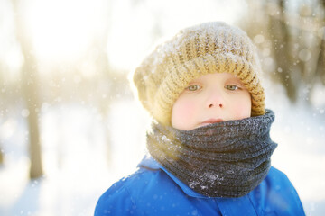 Fototapeta na wymiar Close-up portrait of sad little boy in blue winter clothes walks during a snowfall on cold day. Outdoors winter activities for kids. Cute child wearing a warm clothing, hat and scarf