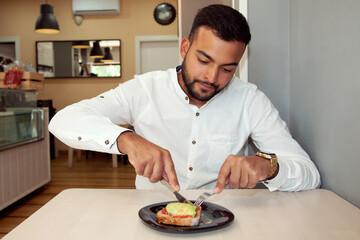 Chico comiendo. Chico en cafetería. Hombre cortando tostada. Tostada de aguacate y tomate. Chico cortando. Hombre comiendo. Lifestyle. Descanso. Hombre con café. Hombre feliz comiendo.  