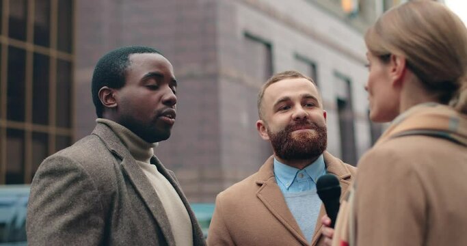 Young successful african american and caucasian businesspeople giving interview to female reporter standing on the street near office building.