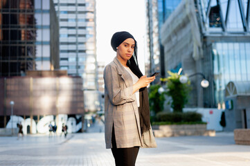 Side view of a muslim woman standing in a urban area looking at camera using her phone