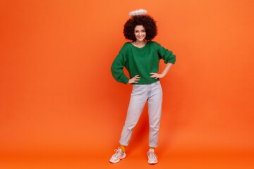 Fototapeta na wymiar Full length of confident angelic woman with Afro hairstyle wearing green casual style sweater standing with hands on hips, having nimb over her head. Indoor studio shot isolated on orange background.