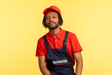 Portrait of bearded handyman with pleasant appearance wearing blue overalls, res T-shirt and visor cap, posing looking directly at camera. Indoor studio shot isolated on yellow background.