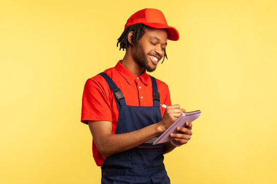 Positive smiling handyman wearing blue overalls and red t shirt writing in notebook, being happy to have lots orders, service industry. Indoor studio shot isolated on yellow background.
