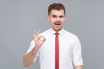 Its Ok. Portrait of young funny handsome man in white shirt and tie standing with Ok sign and looking at camera winking. indoor isolated on gray background.