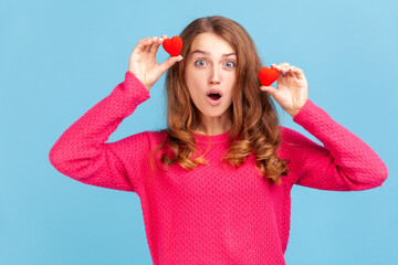 Astonished attractive woman wearing pink pullover, holding two red hearts in hands, looking at camera with open mouth, shocked expression. Indoor studio shot isolated on blue background.
