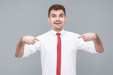 who? me? Portrait of young shocked handsome man in white shirt and tie standing with shocked face, pointing himself and looking at camera with shocked face. indoor isolated on gray background.