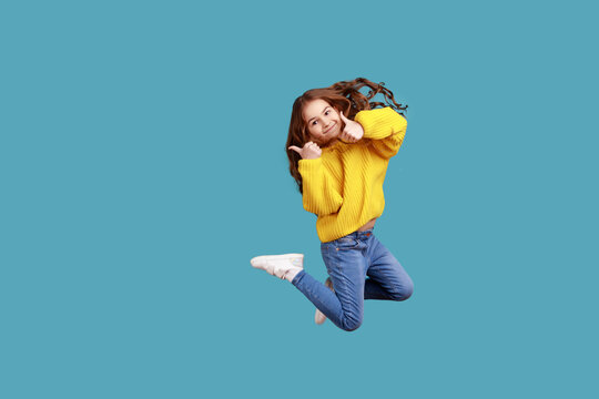 Full length portrait of happy charming little girl jumping up high and showing thumb up to camera, wearing yellow casual style sweater. Indoor studio shot isolated on blue background.