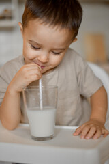 A three-year-old little boy drinks milk from a glass in the kitchen through a paper tube