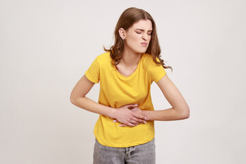 Portrait of unhappy sick teenager girl with brown hair in casual style T-shirt standing, holding her belly with hands, stomach cramps or period pain. Indoor studio shot isolated on gray background.