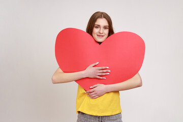 Attractive romantic female of young age holding big read heart and smiling at camera, demonstrating love affection feelings with huge greeting card. Indoor studio shot isolated on gray background.