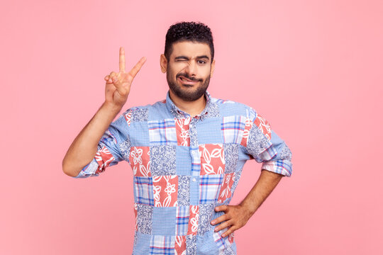 Portrait of excited man winking eye and frowning face in casual blue shirt standing with raised hand and showing v sign or peace, victory gesture. Indoor studio shot isolated on pink background.