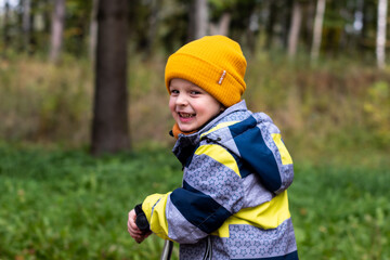 A child rides a scooter in an autumn park. A little boy in a yellow hat rejoices, smiles, laughs, walks in the forest in autumn