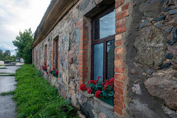 wall with windows of old medieval village house in Latvia. House made of big splitted rocks and clay bricks. Red flowers on windowsills, green grass on ground, reflection of clouds in window glass