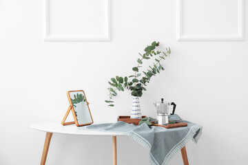 Vase with eucalyptus branches, mirror and coffee maker on table near light wall