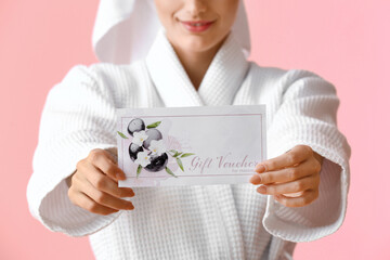 Young woman in bathrobe with gift voucher on pink background, closeup