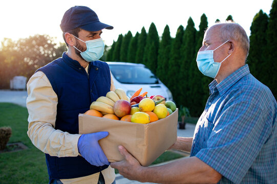 Delivery Man With Protection Mask Delivering Food To The Senior Man During Covid19 Pandemic Or Lockdown.