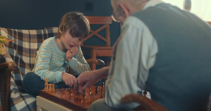 Cute little genius, teenage boy child making smart move playing chess against senior 70s grandfather at living room.