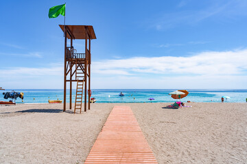 Típica imagen de playa española para vacaciones de sol, deportes y baños recreativos, con una plataforma de acceso hasta el mar y una torre de salvamento de madera con bandera verde. - obrazy, fototapety, plakaty