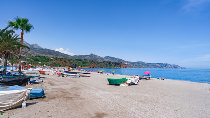 Fototapeta na wymiar Playa con barcas sobre la arena, el mar, acantilados y montañas de fondo un día soleado de vacaciones en verano, con cielo azul. Desde Nerja, Málaga, Andalucía, España.