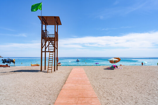 Típica imagen de playa española para vacaciones de sol, deportes y baños recreativos, con una plataforma de acceso hasta el mar y una torre de salvamento de madera con bandera verde.