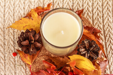 White candle in a glass, red and yellow leaves, cone and  on white knitted background, autumn composition for cozy home atmosphere flat lay