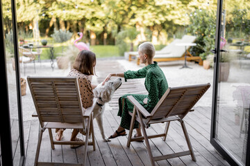 Girlfriends caress dog while sitting on wooden chairs at home terrace. European girls enjoying time together. Concept of modern lifestyle. Idea of female friendship. Sunny warm daytime