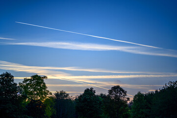 Dark blue sky at dusk with white clouds. An airplane (aeroplane) contrail extends across the sky. Silhouette of trees on the horizon. View of the sky with clouds and contrail just after sunset.