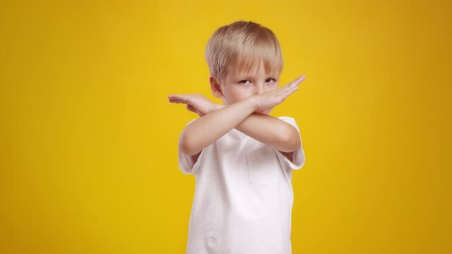 Cute little blonde boy showing stop gesture, crossing hands over orange studio background, slow motion