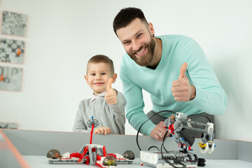 Happy handsome man smiles at the camera with his cheerful little son while playing with Bricks robots and shows the thumbs up.