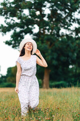 Fototapeta na wymiar Summertime. A happy young woman in a dress and a straw hat posing at the field. In the background is a large tree. Vertical