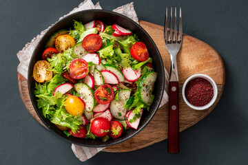 Spring vegetable salad with olive oil and sumac in a black bowl on a dark background. Salad of radishes, cucumbers, tomatoes, lettuce, sumac, and olive oil. Vegan or diet food. Healthy lunch.