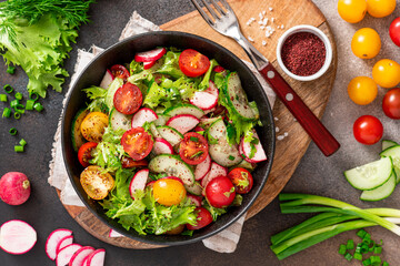 Spring vegetable salad with olive oil and sumac in a black bowl on a dark background. Salad of radishes, cucumbers, tomatoes, lettuce, sumac, and olive oil. Vegan or diet food. Healthy lunch.