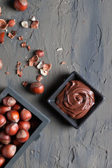 Nutty chocolate paste in a square dish on a dark gray slate, stone or concrete table. Top view. There are a lot of hazelnuts on the table. the concept of sweet food.