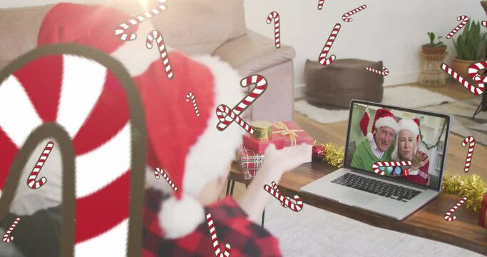 Animation of candy canes, caucasian father and son in santa hats on laptop video call at christmas