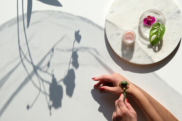 Hand massage, lymph drainage with green jade stone roller. Moisturizing cream on marble plate. Exotic flat lay with monstera leaves, vibrant magenta orchid flowers. Sunlight, long shadows.