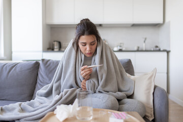 Unhealthy upset young female with fever looking at thermometer with high degree temperature. Sick ill girl suffer from flu or cold, have covid-19 symptoms sitting covered blanket on couch at home. - obrazy, fototapety, plakaty