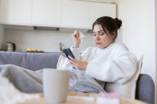 Sick young female with flu browsing, surching possible symptoms on internet by phone, consulting with doctor online on sofa at home. Ill girl with cold chatting with physician in medical app.