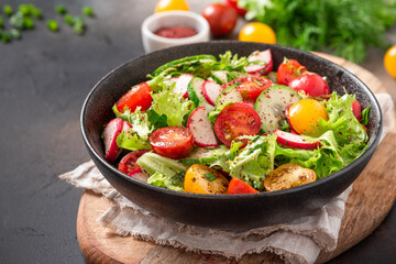 Spring vegetable salad with olive oil and sumac in a black bowl on a dark background. Salad of radishes, cucumbers, tomatoes, lettuce, sumac, and olive oil. Vegan or diet food. Healthy lunch.