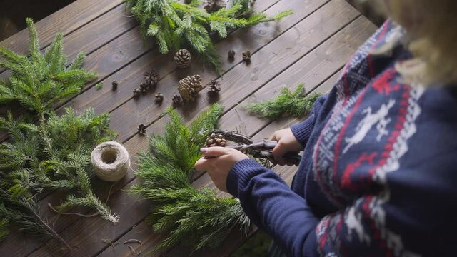 Aged wooden table in authentic florist workshop. Backside view female attache cone on evergreen fir branches arranged in wreath, preparing handmade xmas decoration. DIY and handcraft concept
