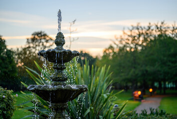 Fountain with water drops in a park with trees behind at dusk. The fountain is seen with a path,...