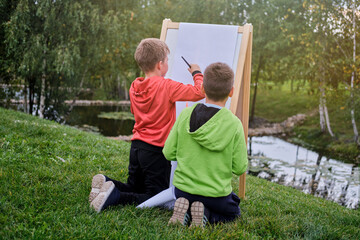 Naklejka premium Children learn to paint on canvas, easel with paper. Boy artist paints nature and trees by the water surface of the river
