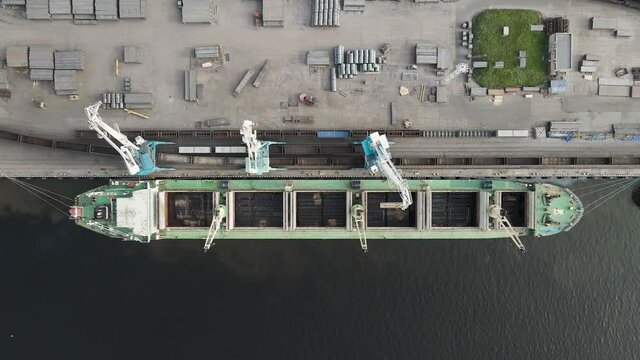 Top down aerial view on a port cranes loading cargo into the hold of the ship. Green ship carrying heavy freight. Vessel is moored at the cargo port. Cargo transportation.